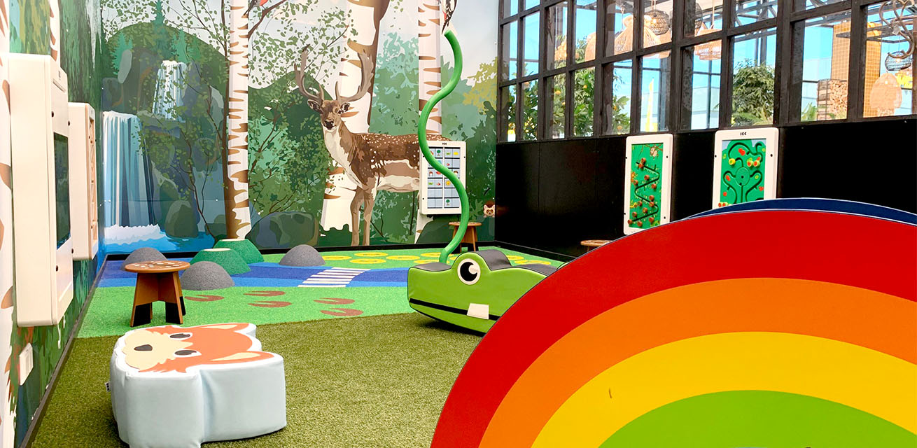 Kinderhoek in een tuincentrum met een glijbaan en meerdere wandspeelborden Kinderhoek in een tuincentrum met een glijbaan en meerdere wandspeelborden en zachte softplay kussens om op te zitten